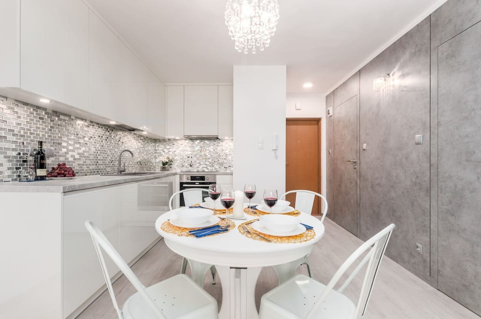 A luxurious dining room with a crystal chandelier, a round dining table set for four with elegant white chairs, and a mirror that adds a sense of space and brightness.