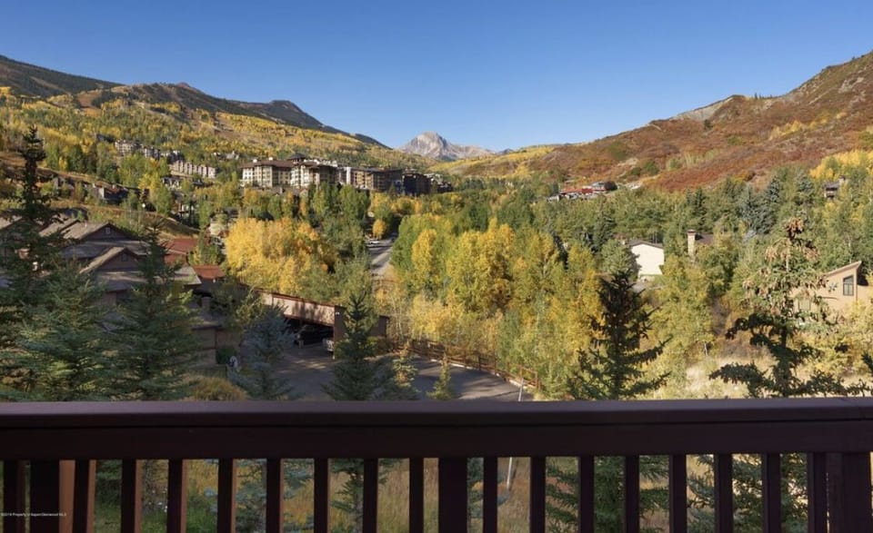 A scenic view from a balcony overlooking a mountainous, autumnal landscape with trees in shades of green and yellow, buildings nestled among trees, and clear blue sky.