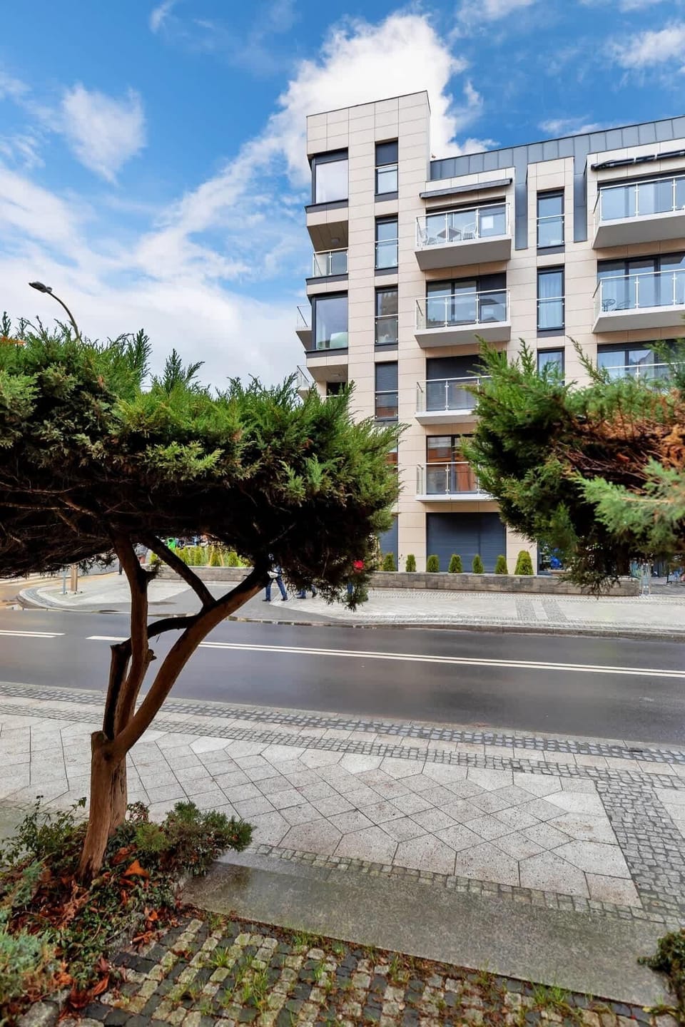 The building features a modern design with white façades, large windows, and a landscaped area with greenery and a paved walkway.