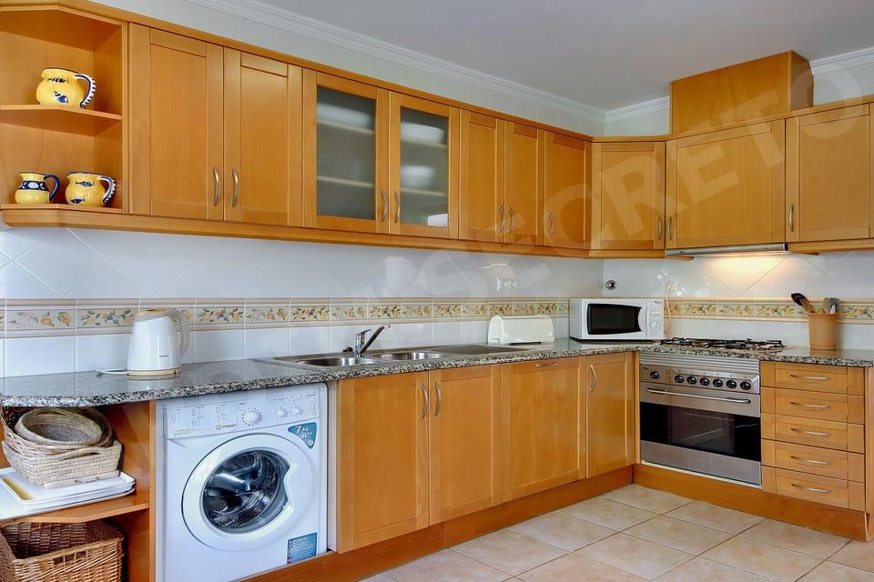 Fully equipped kitchen at Jardim Secreto villa in Carvoeiro, with warm wooden cabinetry, granite worktops, integrated oven and microwave, washing machine, open shelving with decorative ceramics, and tiled flooring