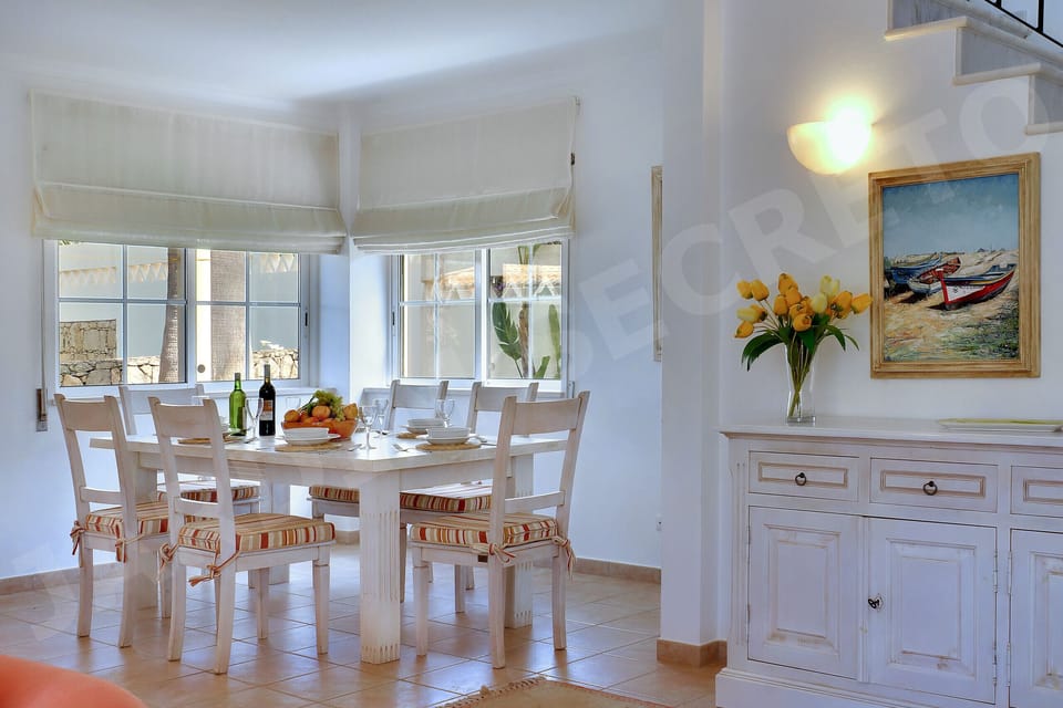 Inviting dining area at Jardim Secreto villa in Carvoeiro, featuring a white dining table with matching chairs, large bay windows flooding the room with natural light and garden views, fresh flowers, a white sideboard, and framed artwork on the walls