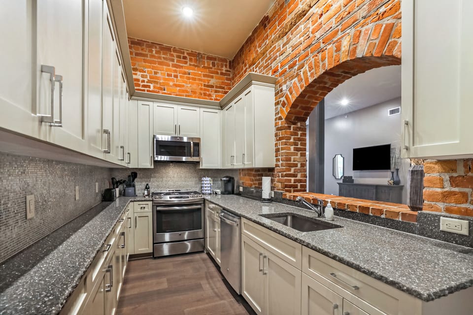 Galley-style kitchen open to living/dining area. Fully stocked with essentials from mixing bowls to wine glasses. Features stainless steel appliances, gas range, stone countertops, and tile backsplash, combining functionality with luxury.