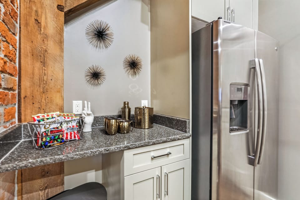 Galley-style kitchen open to living/dining area. Fully stocked with essentials from mixing bowls to wine glasses. Features stainless steel appliances, gas range, stone countertops, and tile backsplash, combining functionality with luxury.