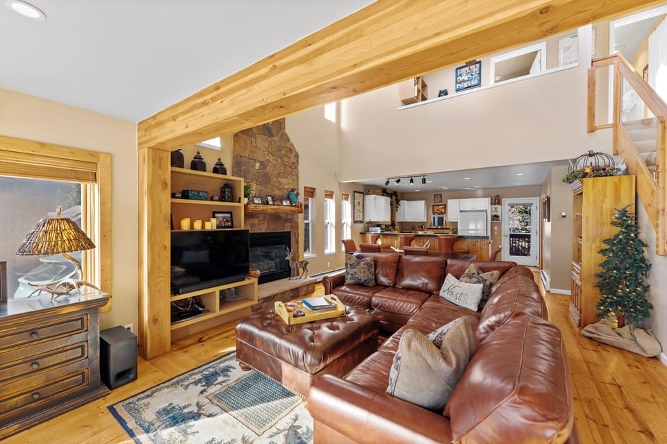 Relax in your spacious living room with rich leather seating, stone fireplace, and exposed wood beams creating mountain cabin charm.