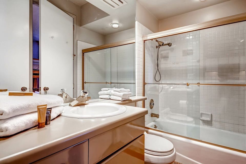 The bathroom has a white vanity and sink, toilet, mirrored wall, and white tiled tub and shower with glass doors.