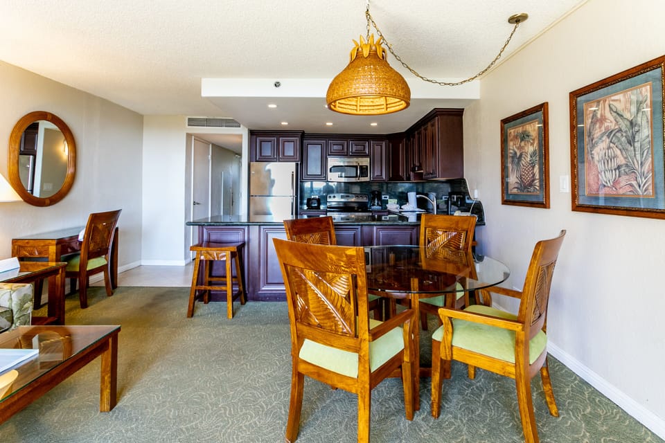 Your island-style dining and kitchen space awaits, featuring rich wood furnishings, a fully equipped kitchen with dark cabinetry, and tropical artwork that sets the perfect vacation mood.
