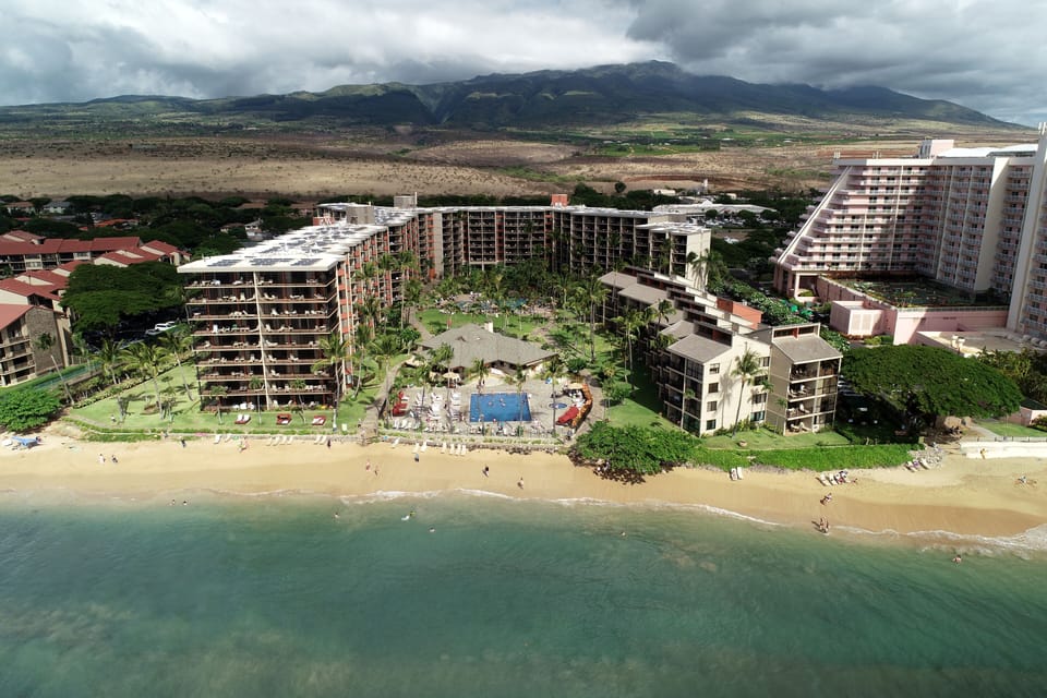 Aerial perspective of a beachfront resort complex nestled between pristine sandy shores and dramatic mountain landscapes, showcasing multiple buildings with ocean access and tropical grounds.