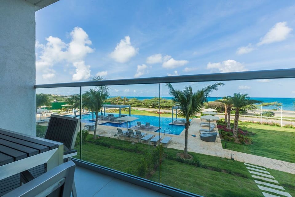A modern beachfront retreat captured from a balcony vantage point. Below, a sparkling pool area is surrounded by lush tropical palms, manicured lawns, and the bright turquoise Caribbean Sea on the horizon.