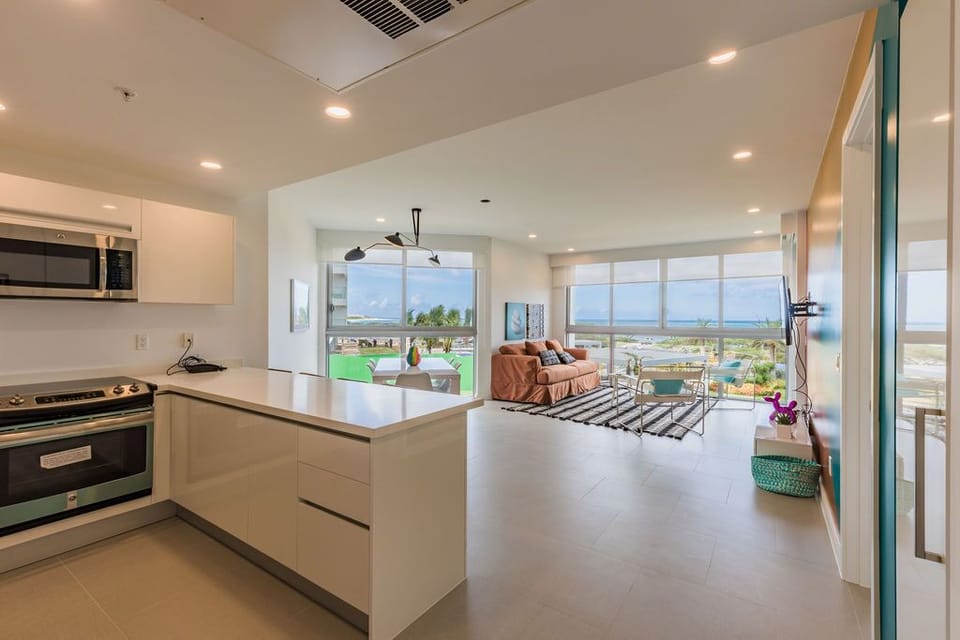 Modern open-concept space with sleek kitchen flowing into a living area, framed by floor-to-ceiling windows showcasing ocean views and tropical greenery beyond.