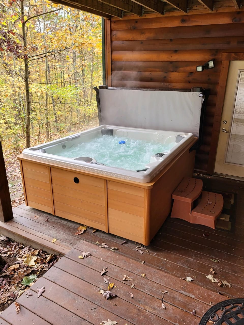 Bubbling hot tub with wooden paneling on a covered porch, surrounded by autumn trees and log cabin walls.