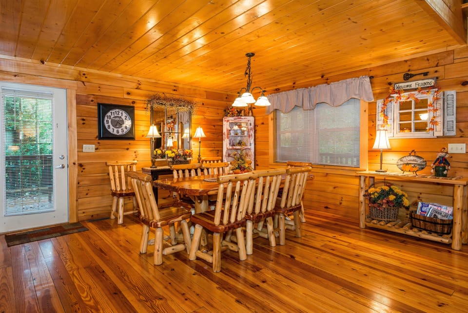 Dining area with polished pine floors, log-style table and chairs, a window with blinds, and decorative shelving.