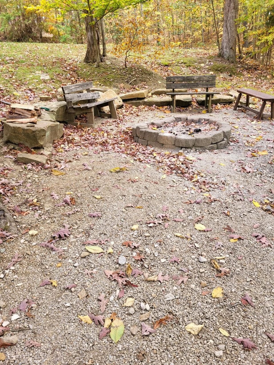 Gravel fire pit area encircled by weathered wooden benches and large natural stone seating, surrounded by autumn leaves.