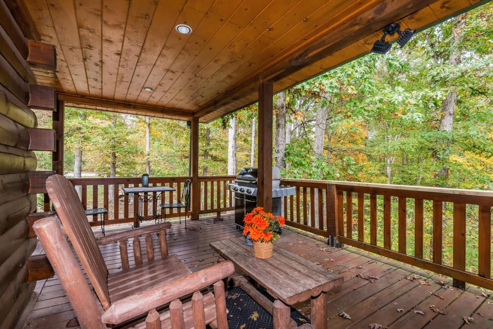Covered wooden deck features rustic Adirondack chairs, a picnic table, gas grill, and forest views.