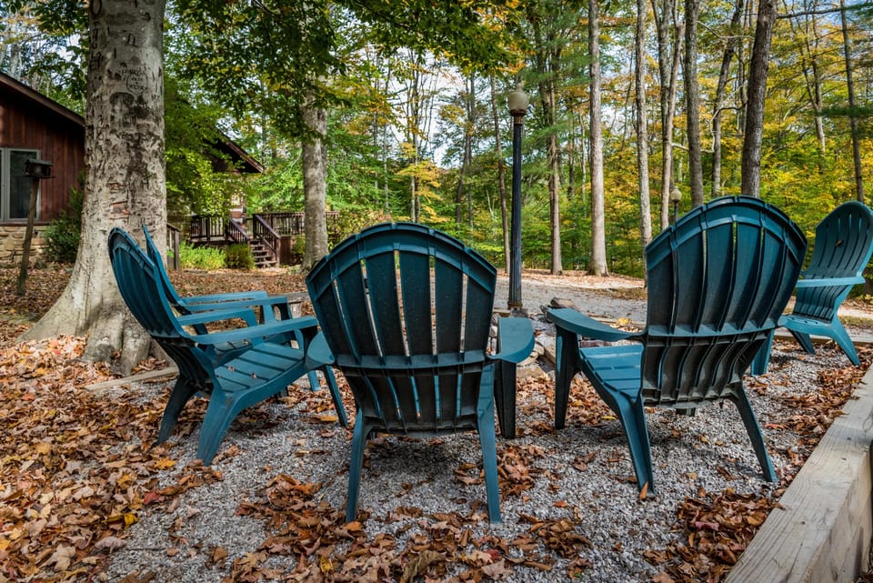 Outdoor seating area with dark teal Adirondack chairs arranged around a fire pit on gravel, surrounded by trees.