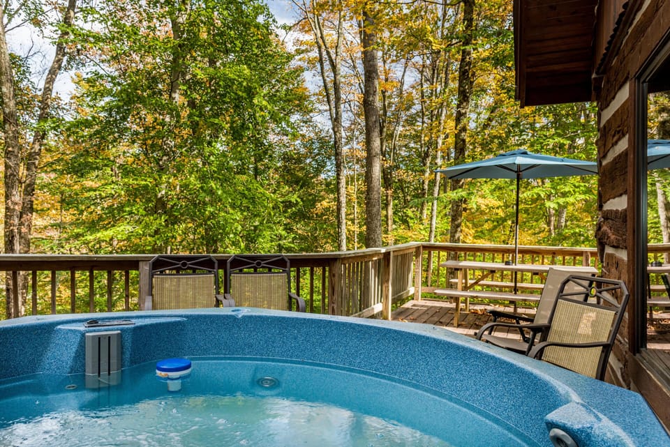 Blue hot tub bubbles on wooden deck with metal chairs and picnic table under a blue umbrella, surrounded by autumn trees.