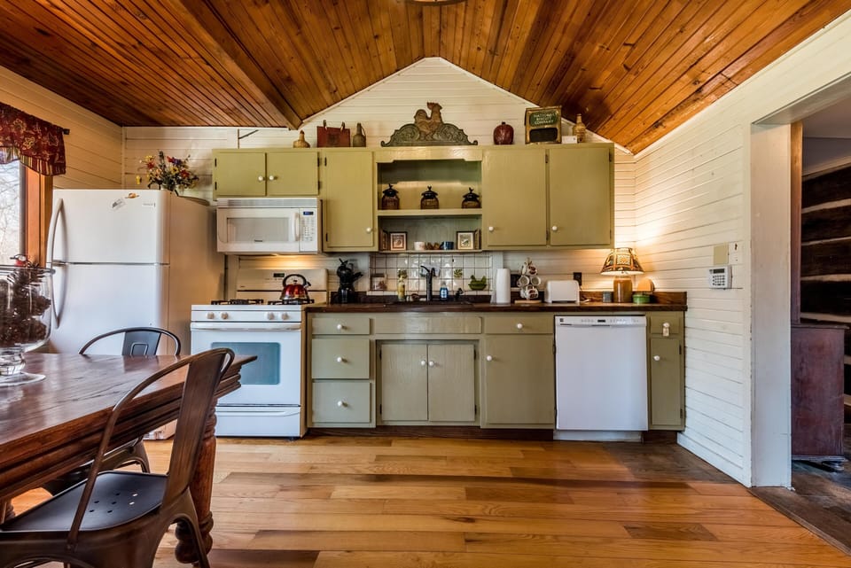 Green cabinets and white appliances line the kitchen wall beneath a vaulted wood ceiling and beside a wooden dining table with metal chairs.