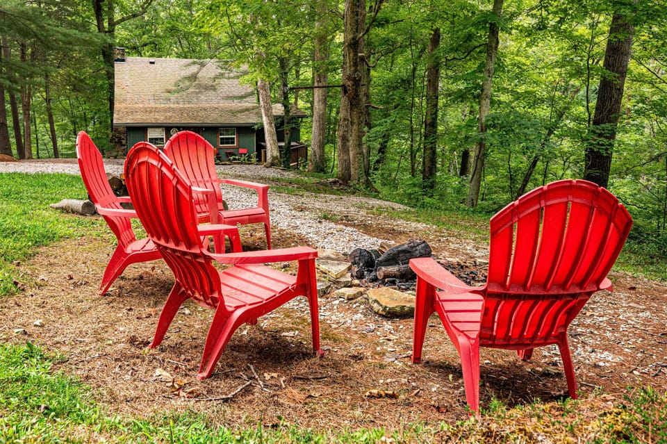 Red Adirondack chairs encircle a stone fire pit with a green wood cabin and forest backdrop.