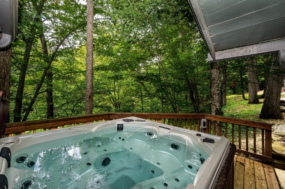 Hot tub on a wooden deck surrounded by dense green trees and a gravel pathway nearby.