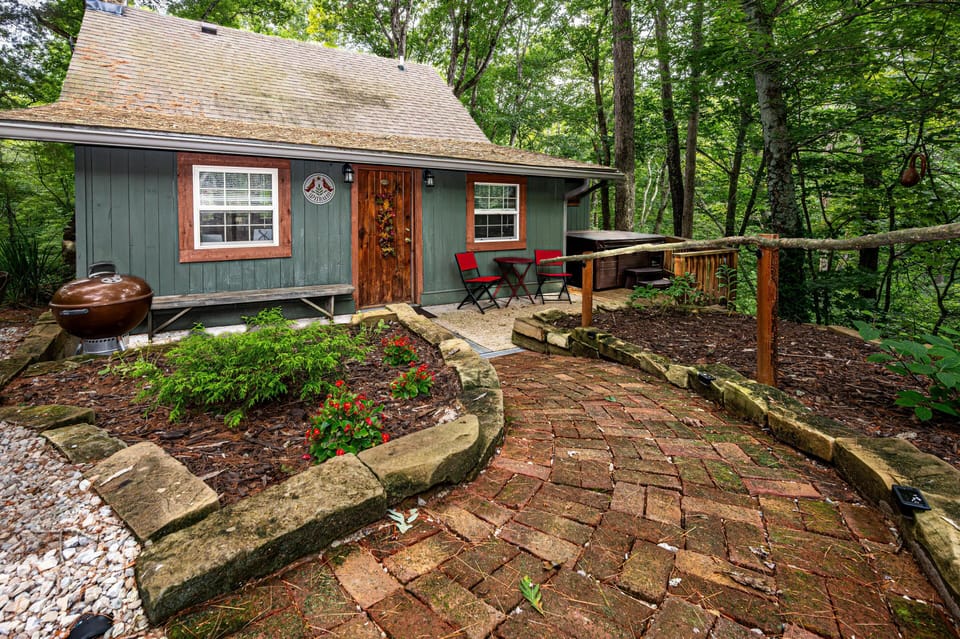 Brick pathway leads to the green wood-sided cabin entrance with red-trimmed windows, a wooden door, and outdoor seating.