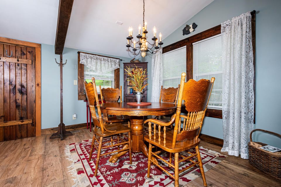 Wooden dining table with carved chairs sits on a red patterned rug beneath a wrought-iron chandelier.