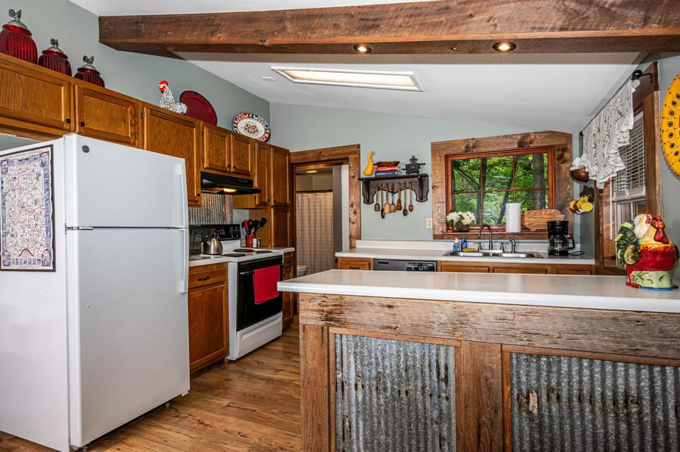 Kitchen with oak cabinetry, white appliances, galvanized metal island panels, and wood-trimmed windows overlooking trees.