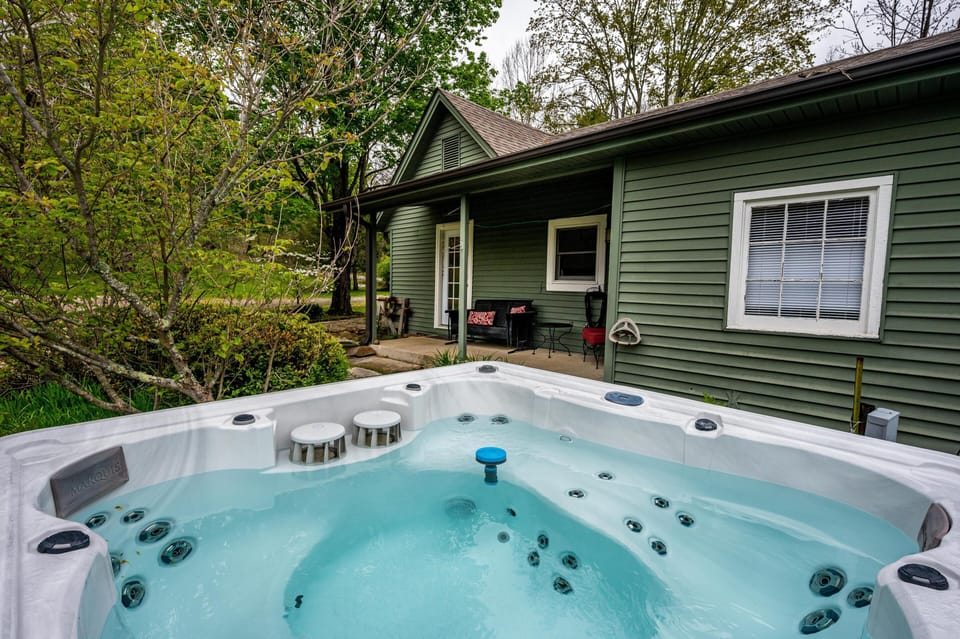 Hot tub with built-in stools sits filled with water beside green wood siding and covered porch seating.