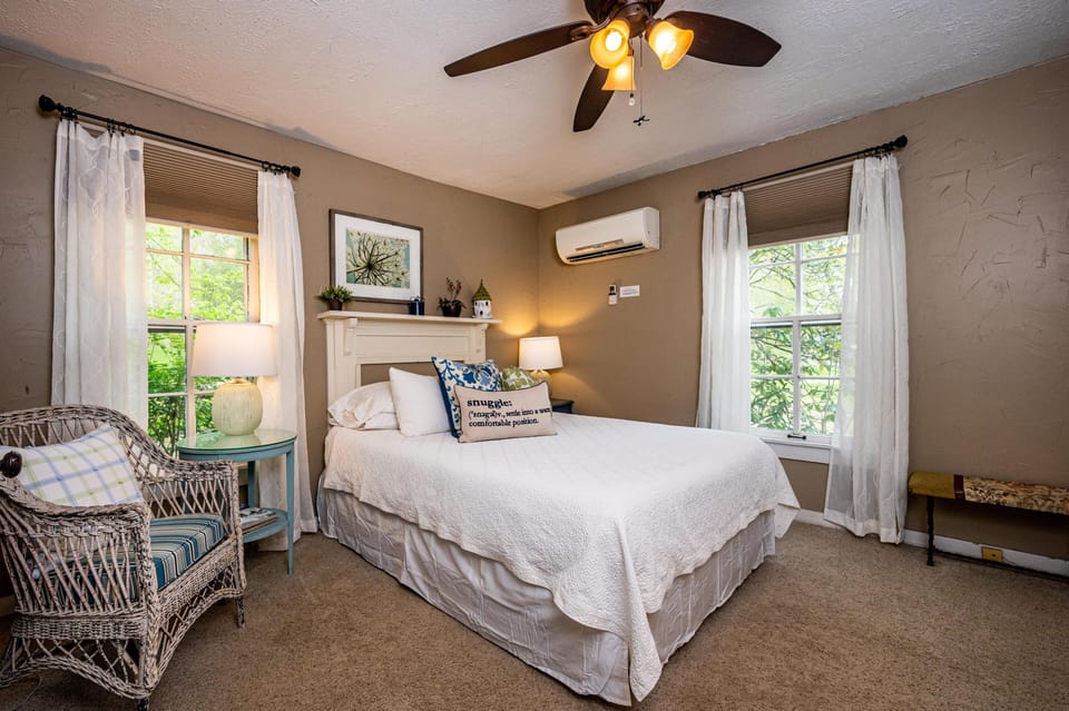 Bedroom with white quilted bedspread, wicker chair with striped cushion, and dual windows with sheer curtains.