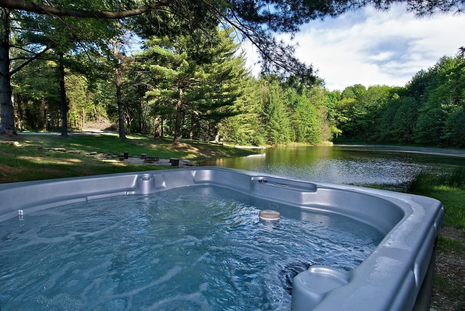 Outdoor hot tub bubbling near the tree-lined private pond with a stone fire pit and grassy shore beyond.
