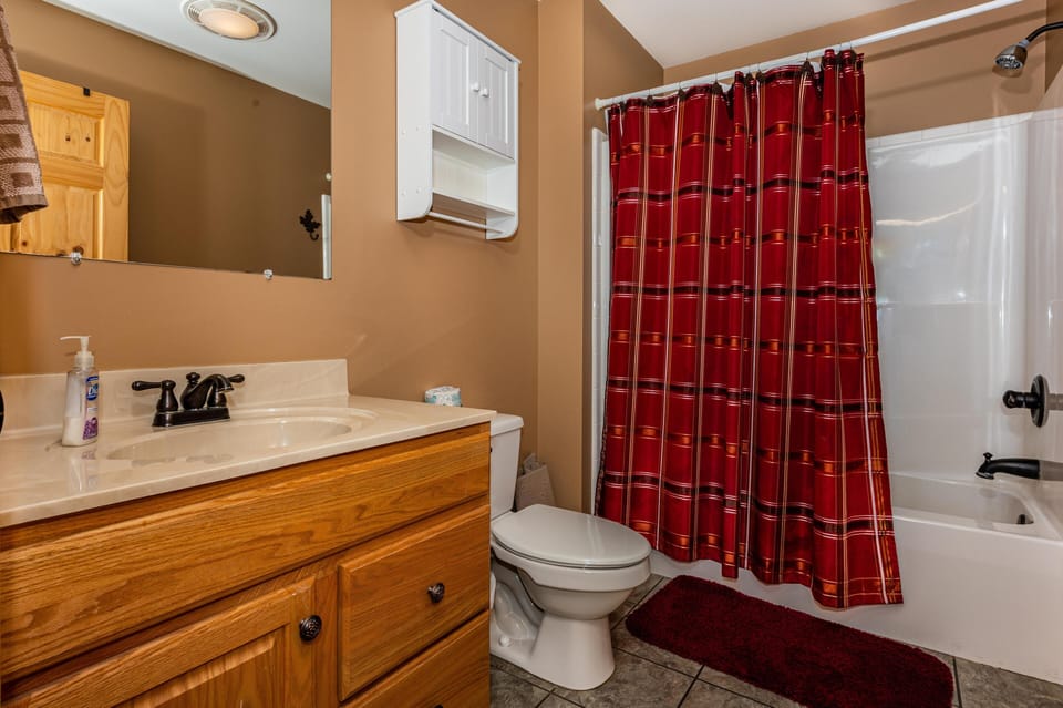 Full bathroom with oak vanity, marble countertop, white bathtub, and red plaid shower curtain on tiled floor.