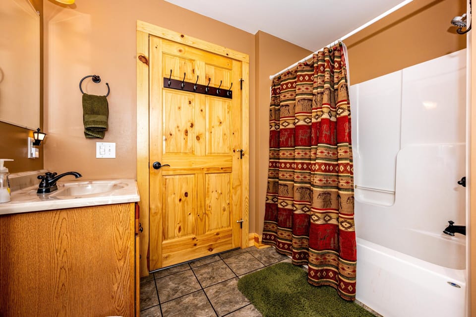 Bathroom with patterned red and brown shower curtain, green rug, pine door, and oak vanity with marble countertop.