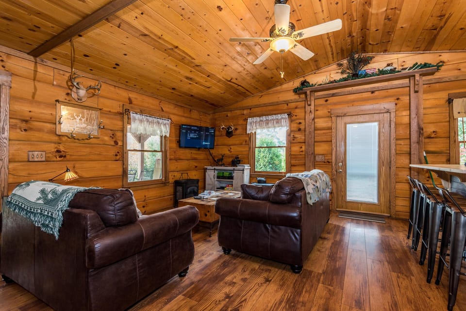 Leather armchairs face a mounted flat-screen TV in the wood-paneled living room with vaulted ceilings and hardwood floors.