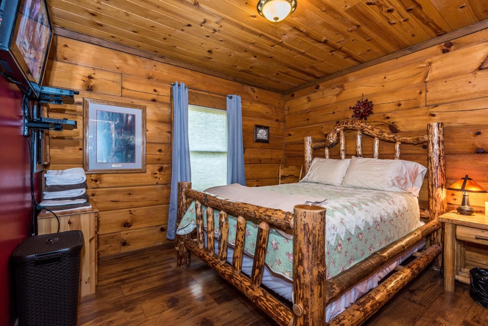 Wood-paneled bedroom with knotty pine walls, hardwood floors, mounted TV, and a nightstand lamp beside the log bed.