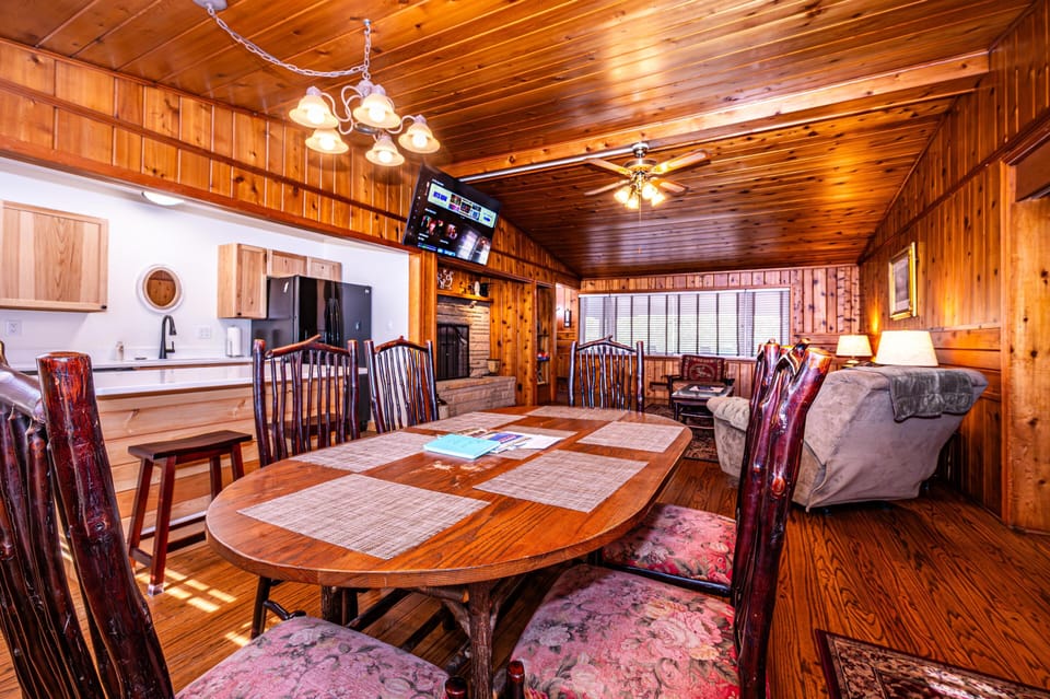 Dining table with floral-upholstered wood chairs sits beside a vaulted knotty pine ceiling and open kitchen with black fridge.
