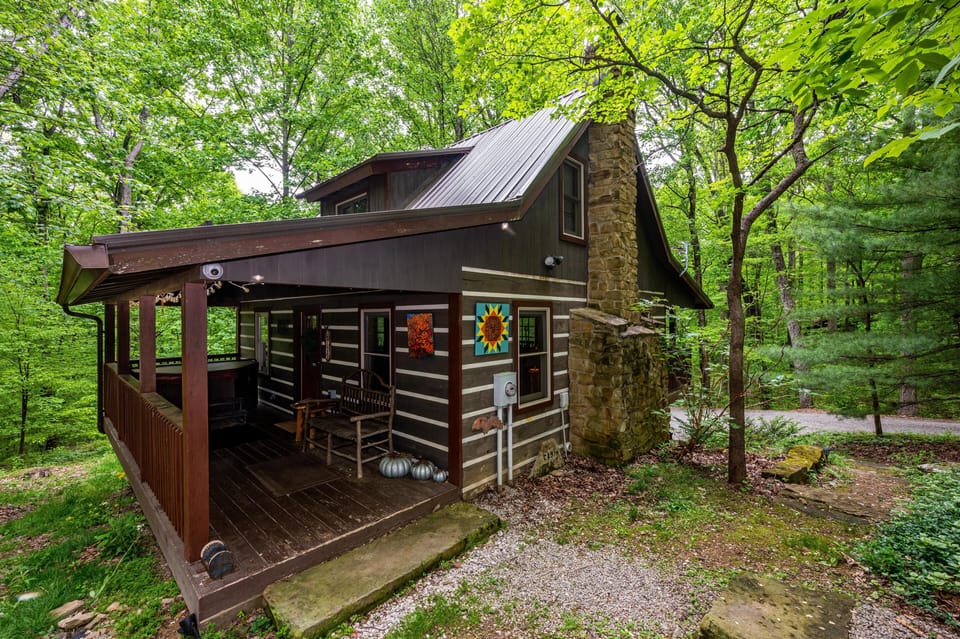 Dark wood cabin exterior with stone chimney and covered porch featuring rustic bench and colorful wall art.
