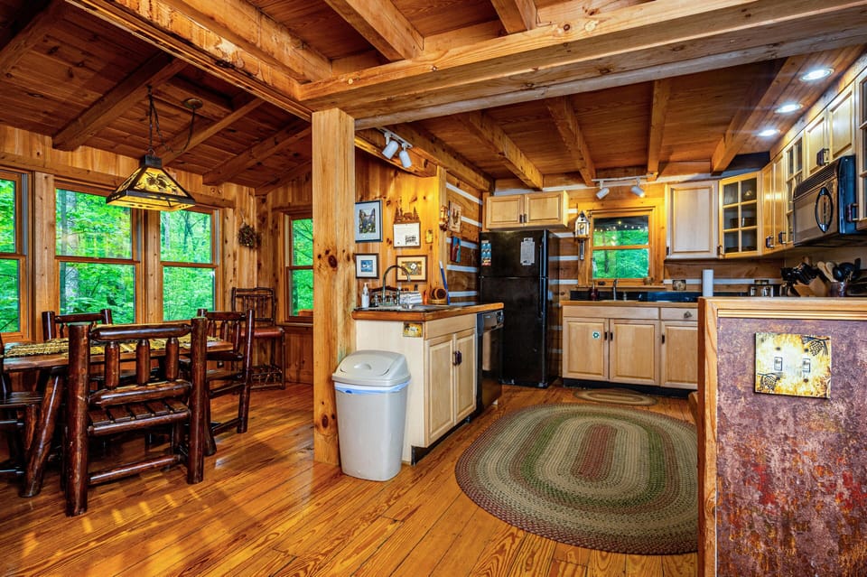 Kitchen and dining area feature honey-toned cabinets, black appliances, a braided oval rug, and large windows with forest views.