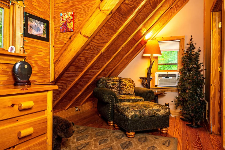 An armchair with matching ottoman, forest-themed upholstery, and wooden legs sits beneath a sloped pine ceiling.