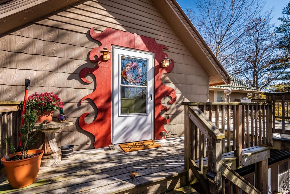 Upper-level wooden deck features a white door framed by red decorative trim, potted plants, and rustic railings.