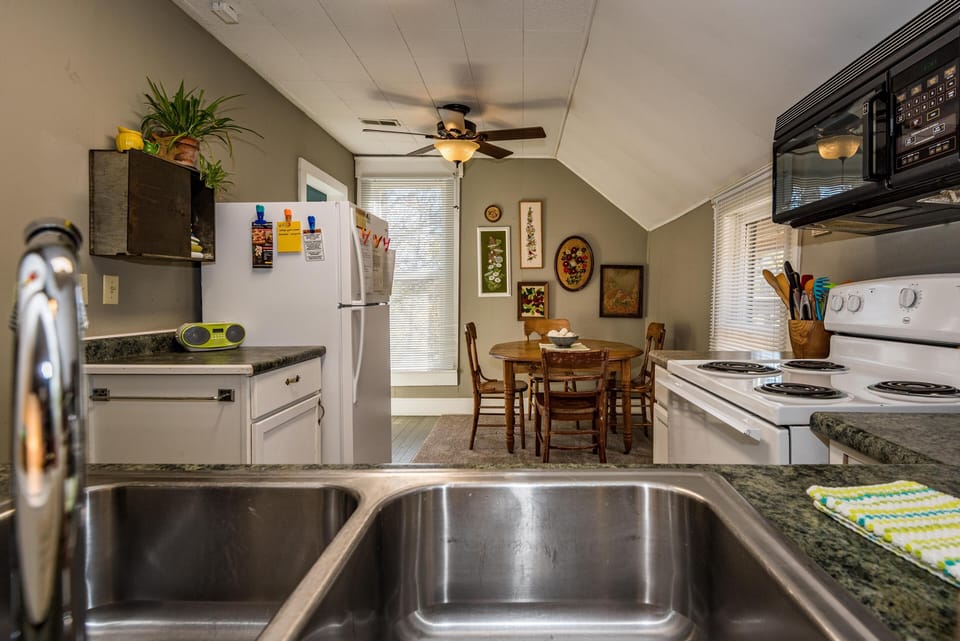 View from stainless steel kitchen sink toward dining area with round wooden table, spindle-back chairs, and floral wall art.