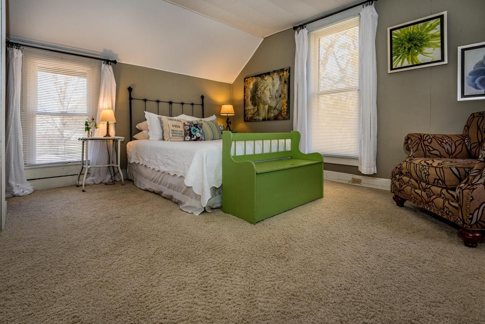 Bedroom with beige carpet, iron bed frame, green wooden bench, floral armchair, and two tall windows.