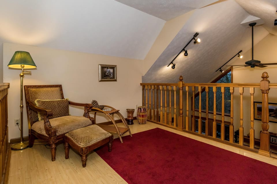 Upstairs nook with patterned armchair, matching ottoman, brass floor lamp, and red area rug by wooden railing.