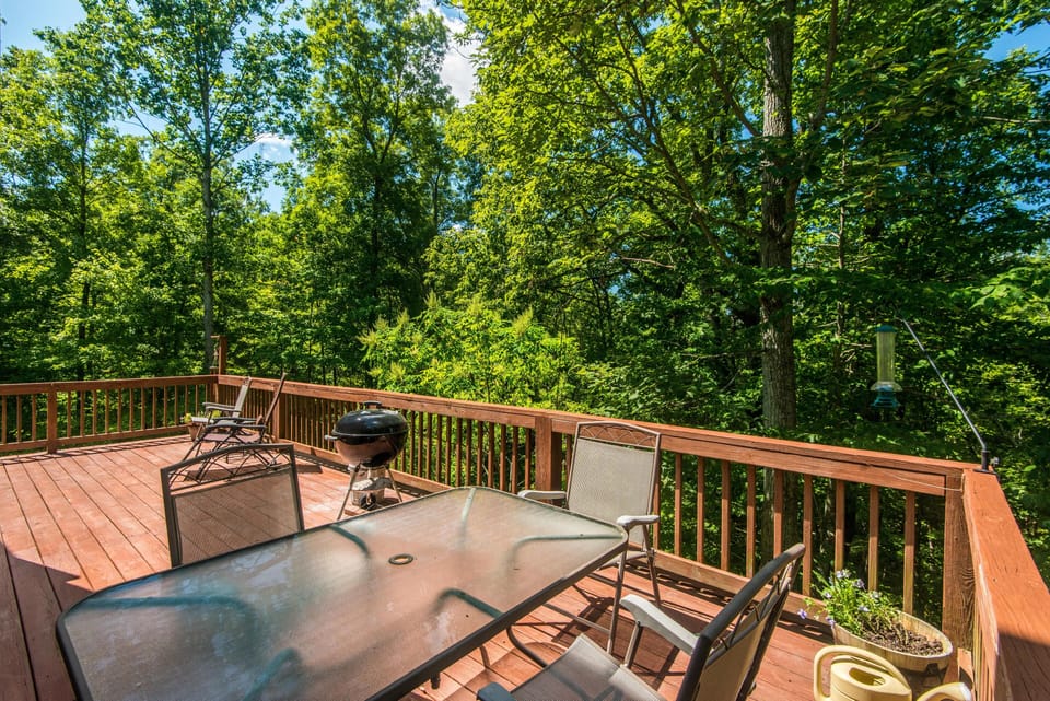 Sunlit wooden deck furnished with a glass-top table, metal chairs, charcoal grill, and forest views.
