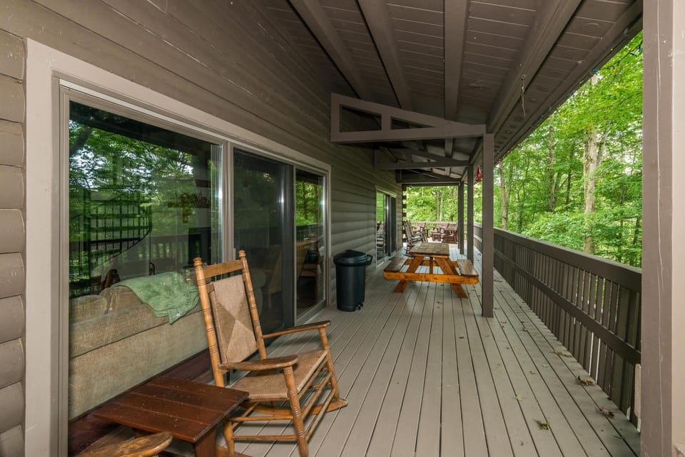 Covered wooden deck with rocking chairs, picnic table, and railing overlooking green forest foliage.