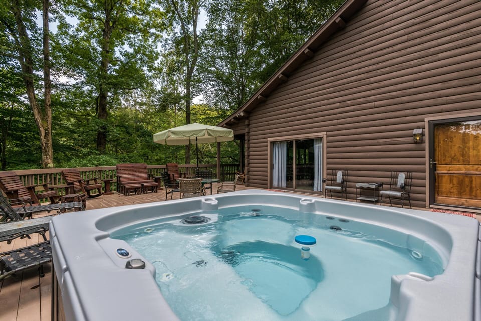 Hot tub bubbling on a wood deck with multiple wooden chairs, table, umbrella, and forest backdrop.