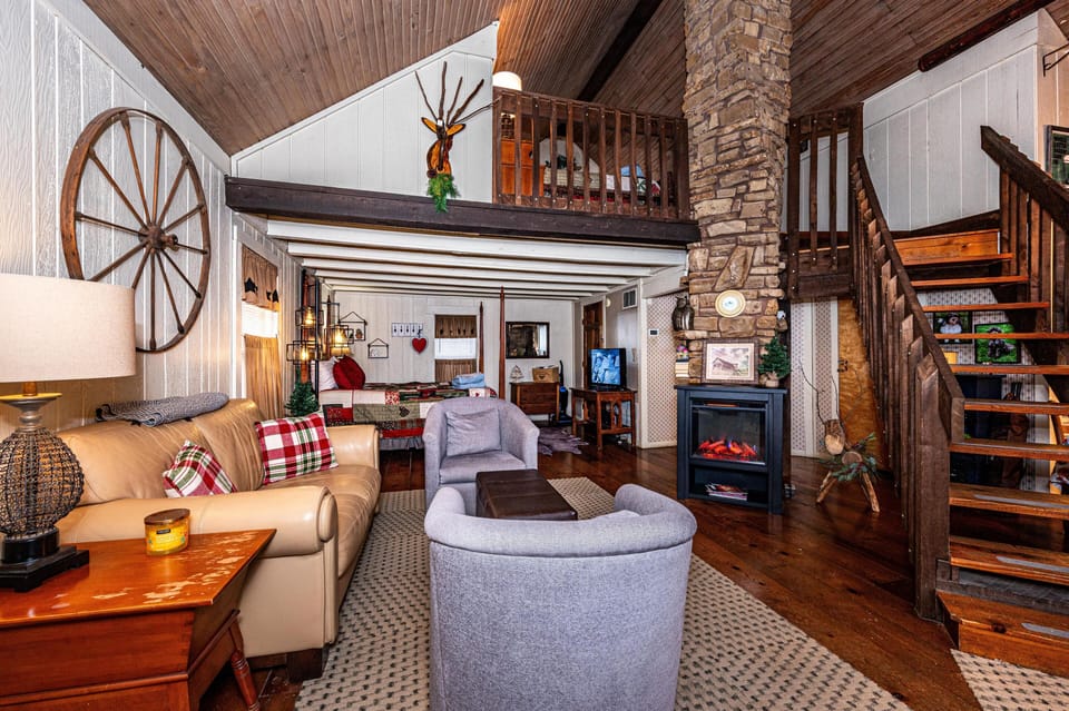 Rustic living area with leather sofa, plaid pillows, stone fireplace, and wood staircase leading to loft bedroom.