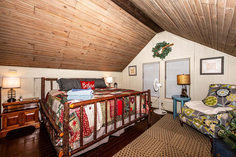Loft bedroom with rustic log bed, patchwork quilt, floral armchair, and slanted wood-paneled ceiling.