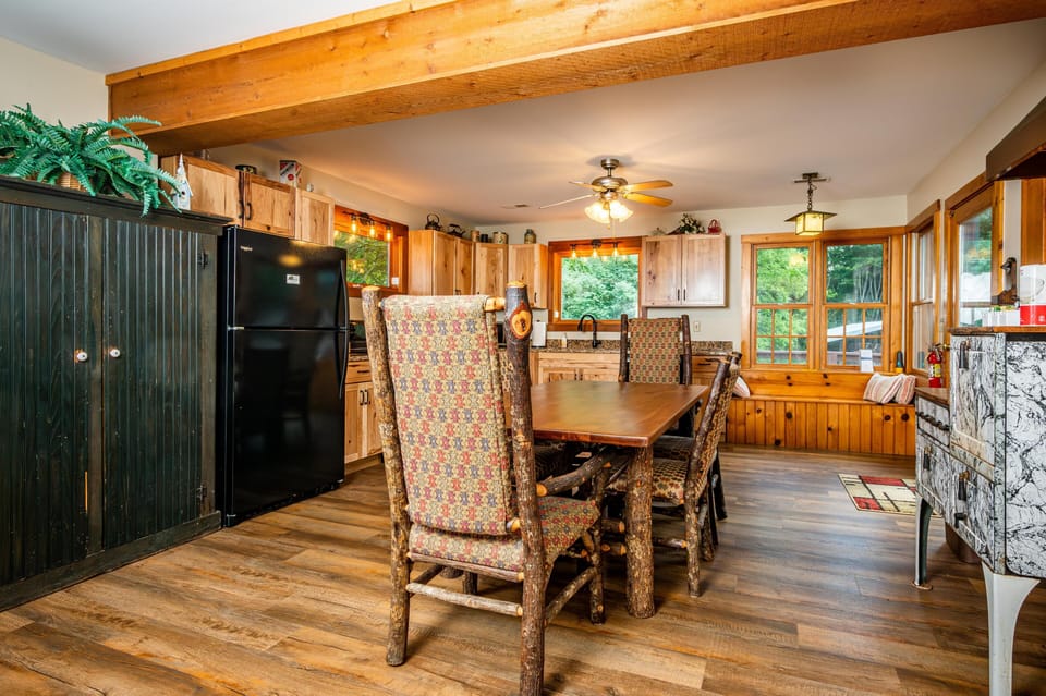 Dining area features a rustic wood table with patterned high-back chairs and large windows overlooking greenery.