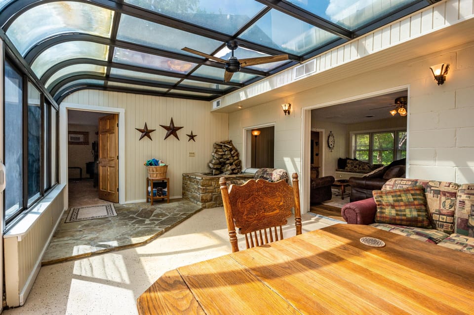 Sunroom with curved glass ceiling, stone fireplace, and wood dining table illuminated by natural light.