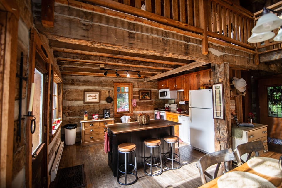 Sunlight illuminates the kitchen island with wood stools, surrounded by exposed log walls and rustic cabinetry.