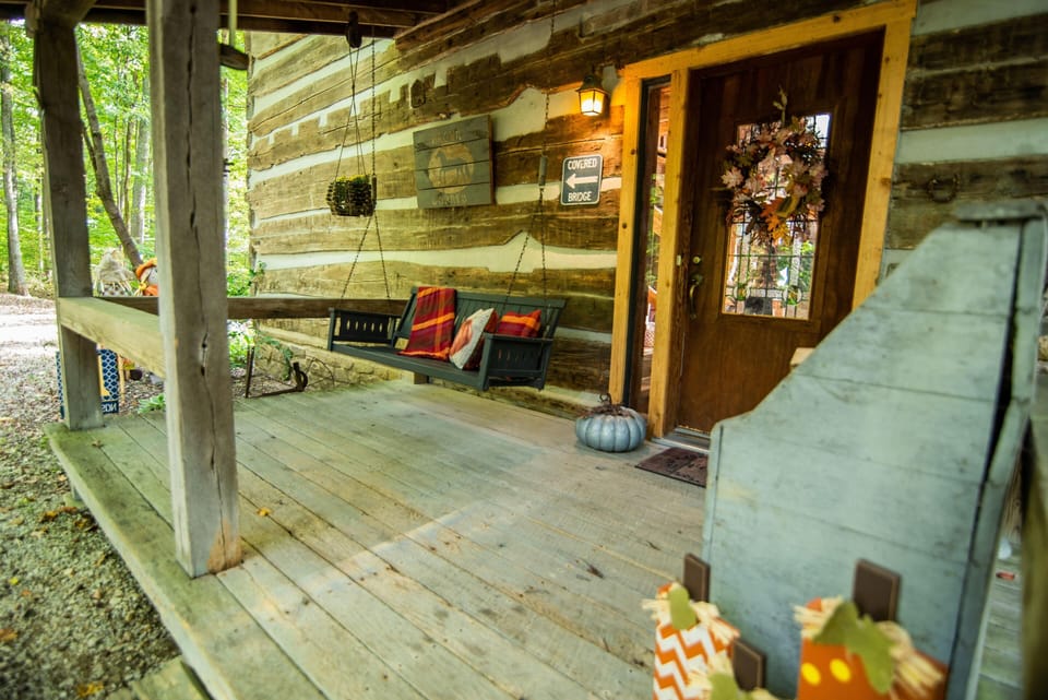 Front porch with weathered wood planks, a dark green hanging bench adorned with red plaid pillows, and a wooden entry door decorated with a fall wreath.