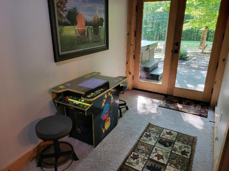 Retro arcade game table with stools sits on carpet near glass doors opening to wooded backyard.